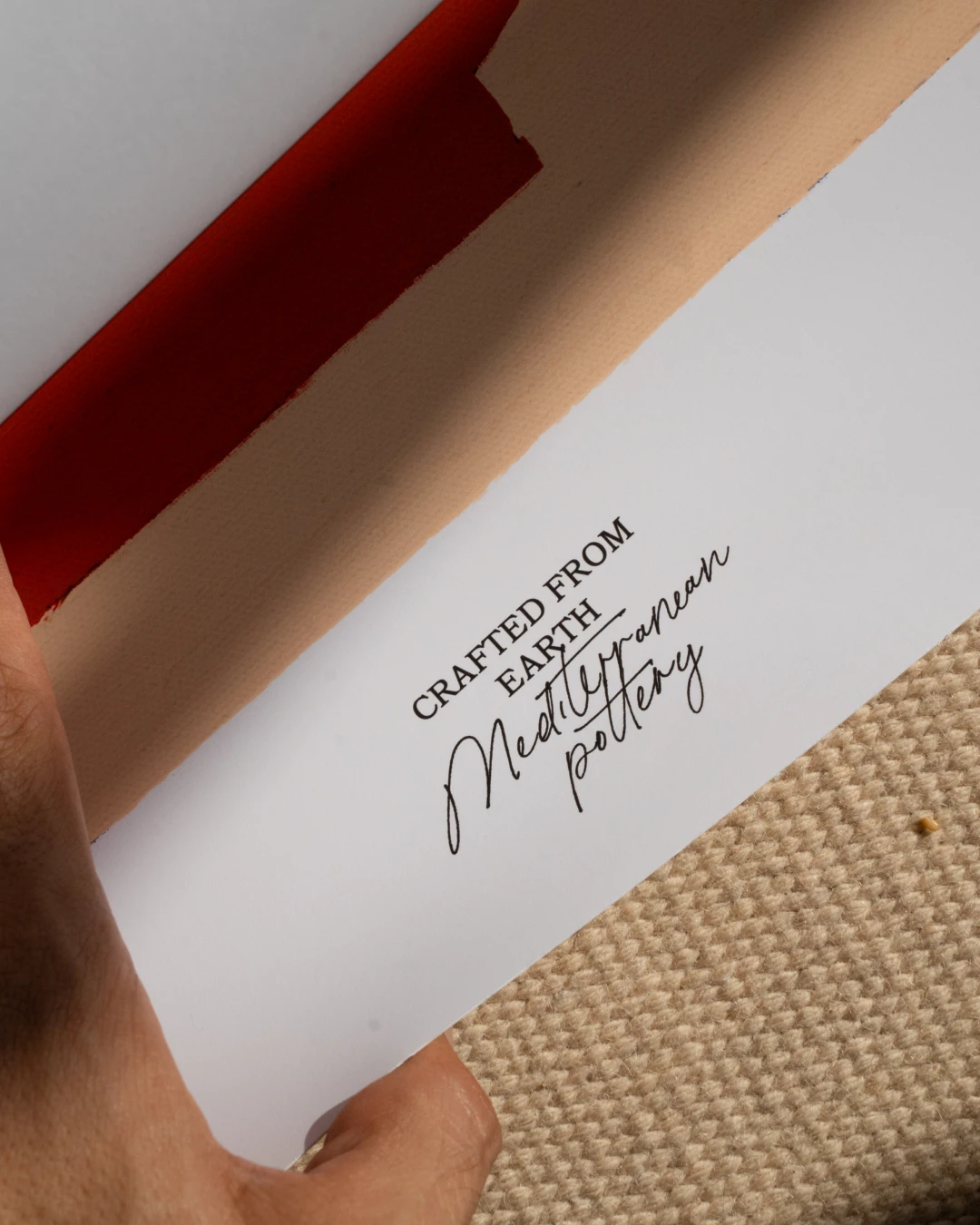 all-groups - A hand holding the white border of the Stamnos print, printed with "Crafted from Earth, Mediterranean pottery", with the red and peach artwork behind it and beige woven fabric below.