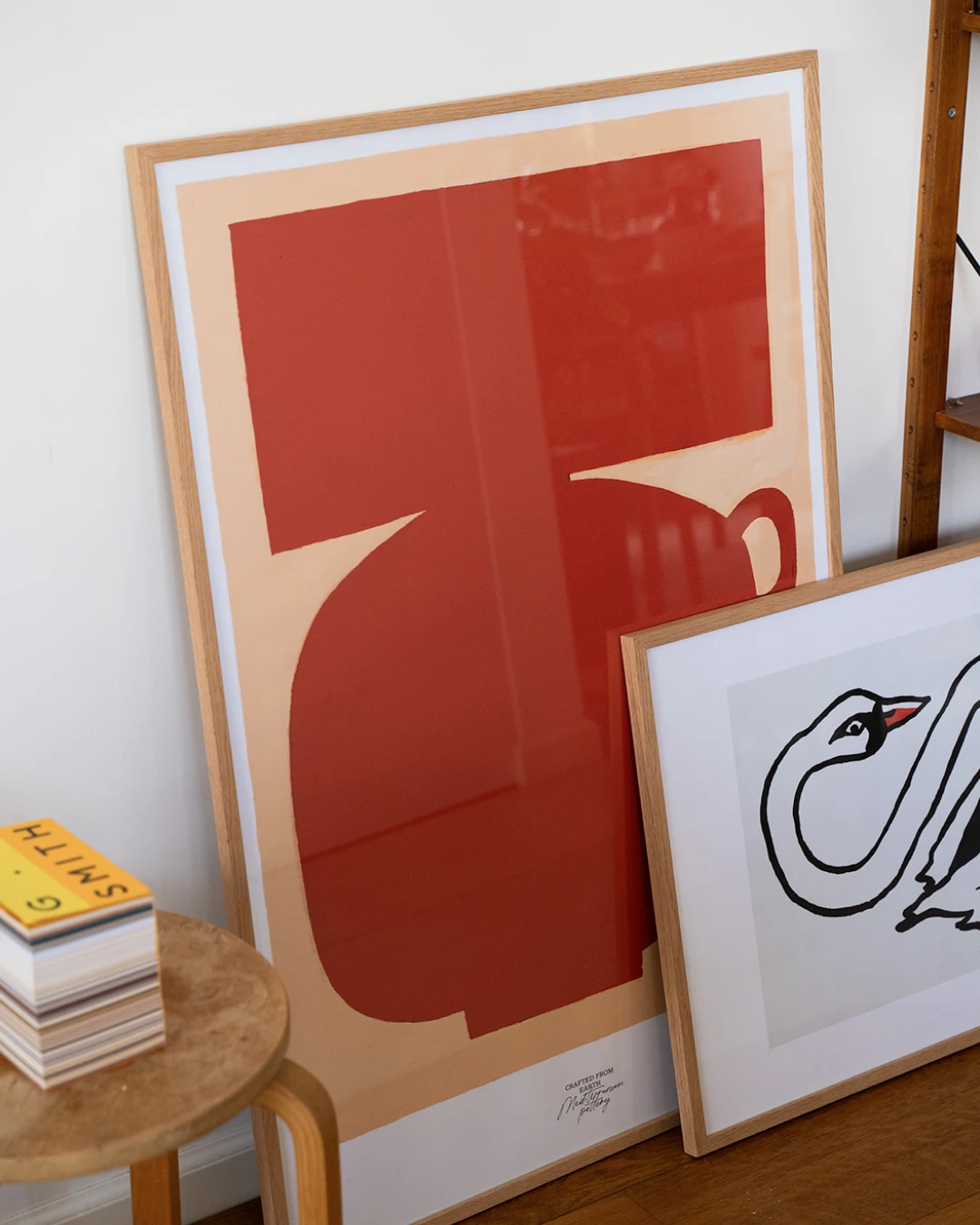 all-groups - Oak-framed Stamnos print leaning against a white wall beside a smaller oak-framed Swan print, with a stack of books on a round wooden stool and a wooden shelf at the top right.
