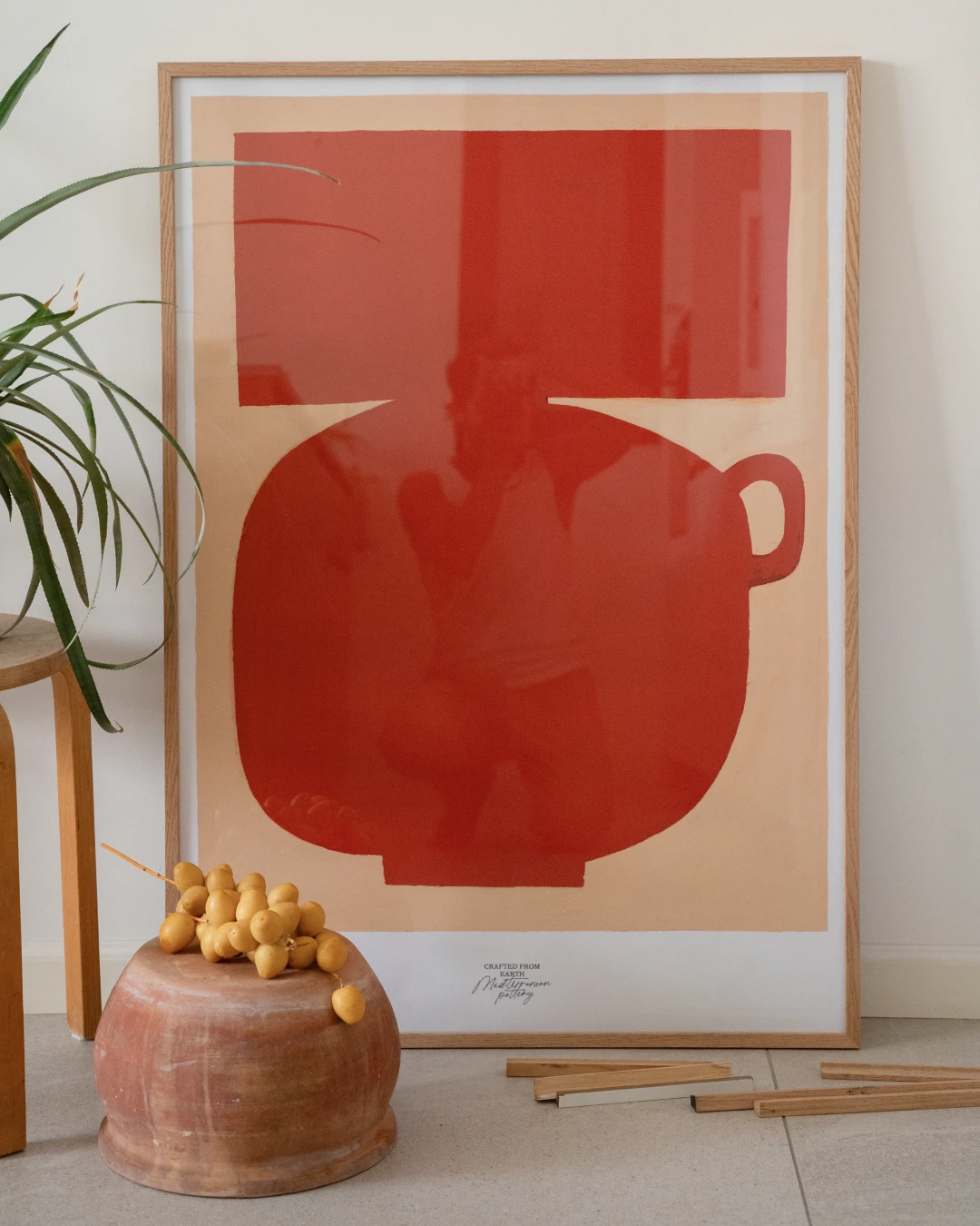 all-groups - Oak-framed Stamnos print leaning against a white wall, with a terracotta pot holding a bunch of small yellow fruit on the floor, a green plant and wooden stool on the left, and small wooden frame pieces on the floor.