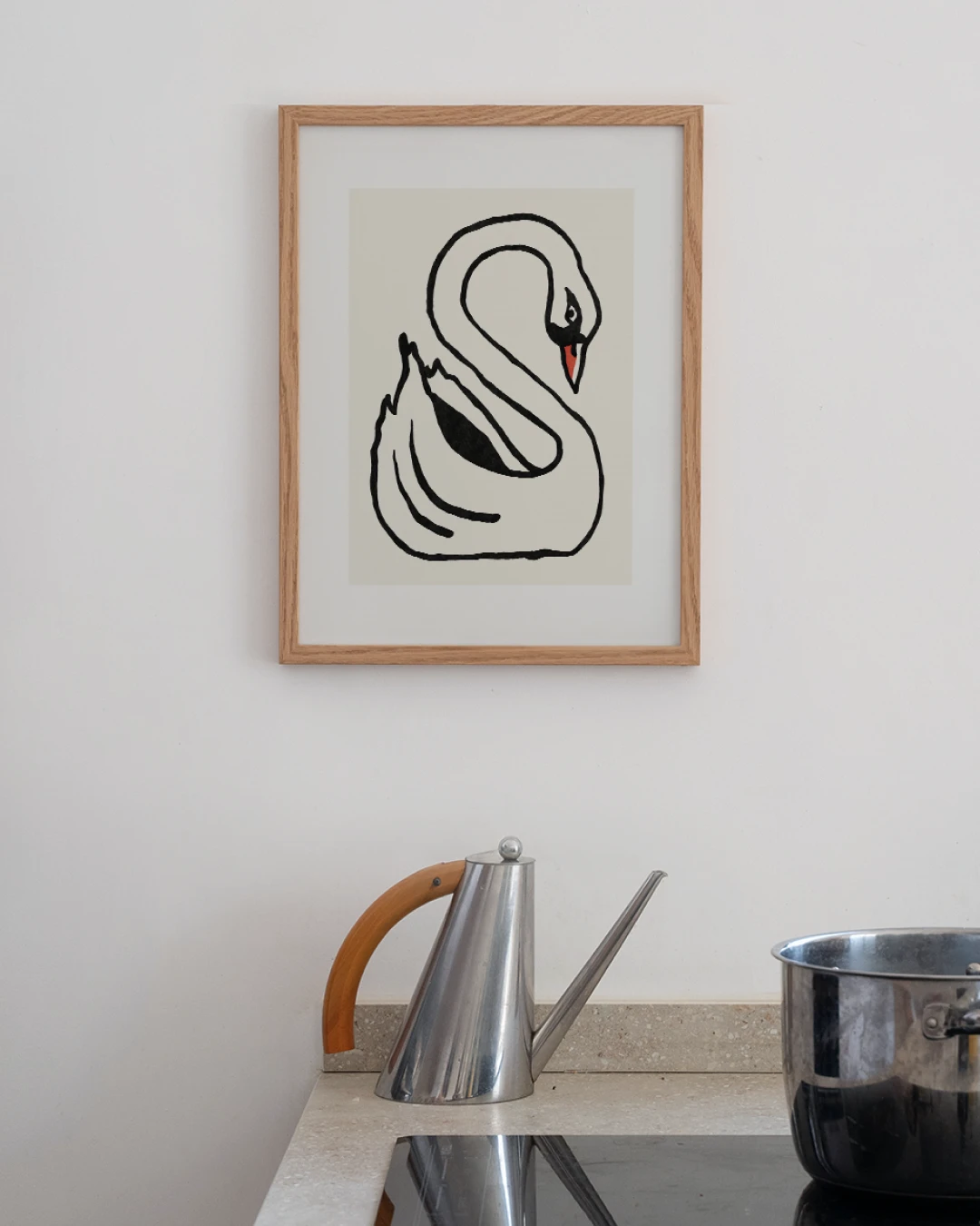 all-groups - Small oak-framed Swan print hanging on a white wall above a kitchen counter, with a steel jug with a wooden handle and a steel pot on the hob below.