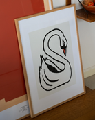 all-groups - Oak-framed Swan print leaning against a wall on a wooden floor, in front of a larger red and peach print, with a wooden shelf and a bowl of tomatoes at the top right.